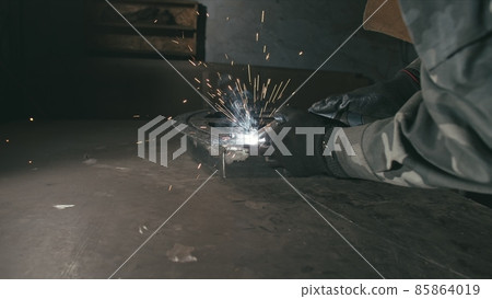 Forge workshop. Smithy. Worker in a welding hood helmet welds a part by electric welding. Sparks are reflected in the protective screen. Forge workshop. Smithy. Worker in a welding hood helmet welds a part by electric welding. Sparks are reflected in the protective screen. 85864019