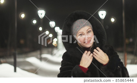 Pretty woman walking along the black fur coat. Outdoor. Girl walks on a winter night among the street lights of the city. Snow park with garlands. 85865444