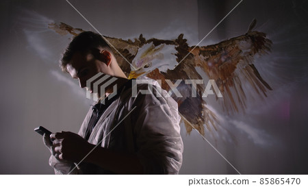 Artist designer draws an eagle on wall. Craftsman decorator paints picture with acrylic oil color looking at sketch in phone. Dark magic cinematic. 85865470