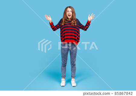 Full length portrait of surprised woman opening her mouth and raised arms in amazement, hearing awesome news, pleasantly surprised by sudden success. Indoor studio shot isolated on blue background. 85867842