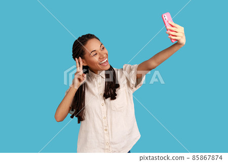 Glad woman with toothy smile, makes peace sign or V gesture at camera of cell phone, poses for making selfie or video call, wearing white shirt. Indoor studio shot isolated on blue background. Glad woman with toothy smile, makes peace sign or V gesture at camera of cell phone, poses for making selfie or video call, wearing white shirt. Indoor studio shot isolated on blue background. 85867874