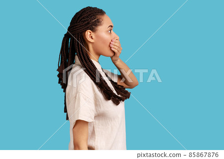 Side view of woman with dreadlocks wants to scream, covers mouth with palm, stares at something terrible, has eyes popped out, wearing white shirt. Indoor studio shot isolated on blue background. Side view of woman with dreadlocks wants to scream, covers mouth with palm, stares at something terrible, has eyes popped out, wearing white shirt. Indoor studio shot isolated on blue background. 85867876