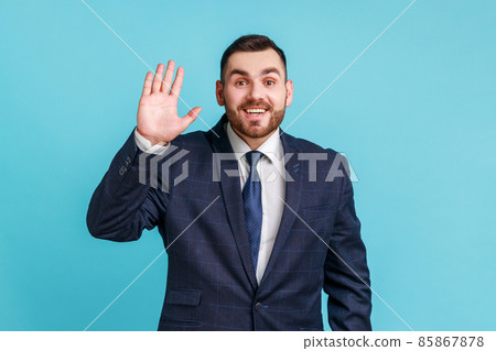 Hello. Portrait of friendly glad handsome brunette man with beard wearing official style suit showing hi gesture with waving hand and smiling sincerely Indoor studio shot isolated on blue background. Hello. Portrait of friendly glad handsome brunette man with beard wearing official style suit showing hi gesture with waving hand and smiling sincerely Indoor studio shot isolated on blue background. 85867878