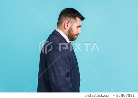 Side view of sad handsome businessman wearing dark official style suit standing with bowed head, having problems at work or with girlfriend. Indoor studio shot isolated on blue background. 85867881