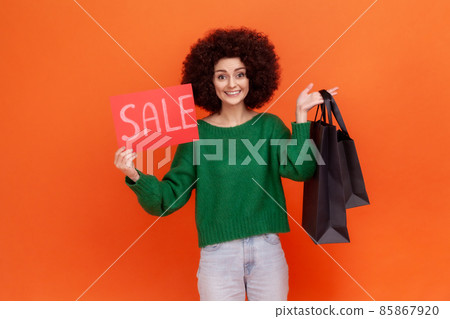 Happy satisfied woman with Afro hairstyle wearing green casual style sweater holding card with sale inscription and black shopping bags. Indoor studio shot isolated on orange background. 85867920