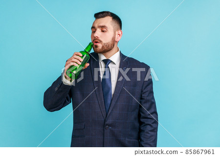 Bearded young adult businessman wearing official style suit drinking alcohol, holding beer bottle in hand, suffering from alcoholism. Indoor studio shot isolated on blue background. 85867961