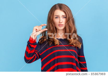 Too small. Portrait of dissatisfied woman in striped casual sweater showing a little bit gesture, inch or centimeter, disappointed with minimum size. Indoor studio shot isolated on blue background. Too small. Portrait of dissatisfied woman in striped casual sweater showing a little bit gesture, inch or centimeter, disappointed with minimum size. Indoor studio shot isolated on blue background. 85867969