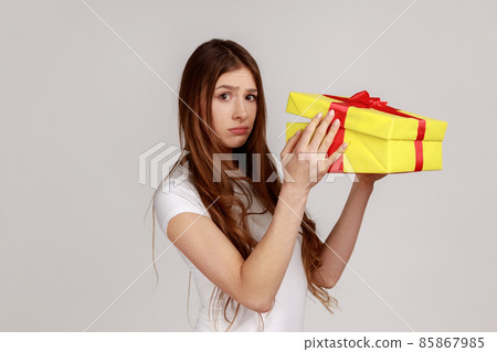 Portrait of upset displeased woman looking at camera, holding gift box, opening present with dissatisfied expression, wearing white T-shirt. Indoor studio shot isolated on gray background. 85867985