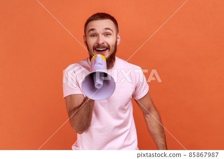 Portrait of optimistic bearded man screaming in megaphone, announcing important information with positive, looking at camera, wearing pink T-shirt. Indoor studio shot isolated on orange background. Portrait of optimistic bearded man screaming in megaphone, announcing important information with positive, looking at camera, wearing pink T-shirt. Indoor studio shot isolated on orange background. 85867987