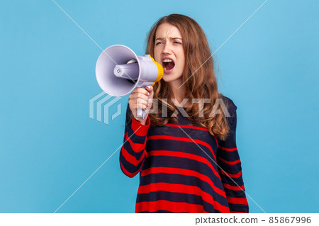 Portrait of woman wearing striped casual style sweater, screaming loudly in megaphone, announcing important advertisement, protesting. Indoor studio shot isolated on blue background. 85867996