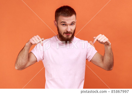 Portrait of man pointing fingers down paying attention at advertising area, showing freespace, looking at camera, wearing pink T-shirt. Indoor studio shot isolated on orange background. 85867997