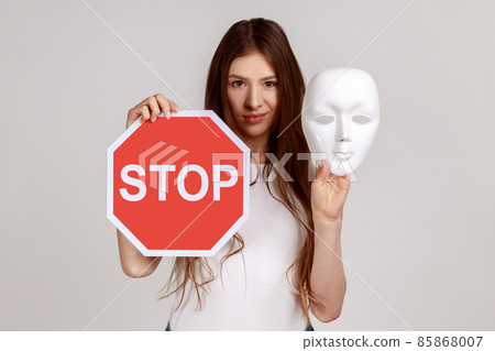 Portrait of serious dark haired woman holding white mask with unknown face and red traffic sign, looking at camera, wearing white T-shirt. Indoor studio shot isolated on gray background. 85868007