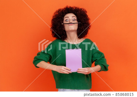 Funny young adult woman with Afro hairstyle wearing green sweater having fun holding pencil with lip and nose, female student resting during hometask. Indoor studio shot isolated on orange background. 85868015