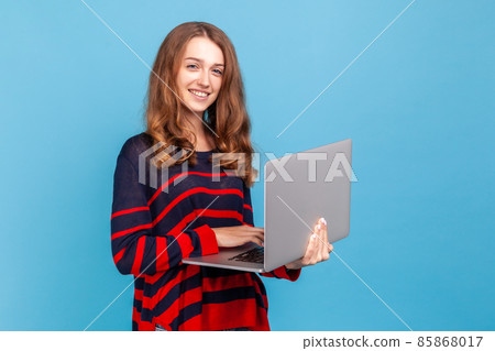 Happy attractive woman wearing striped casual style sweater, standing with laptop in hands and looking at camera with smile, freelancer working online. Indoor studio shot isolated on blue background. Happy attractive woman wearing striped casual style sweater, standing with laptop in hands and looking at camera with smile, freelancer working online. Indoor studio shot isolated on blue background. 85868017