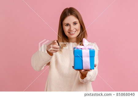 Satisfied young adult blond woman standing with blue gift box in hands and pointing finger to camera, choosing you as winner, wearing white sweater. Indoor studio shot isolated on pink background. Satisfied young adult blond woman standing with blue gift box in hands and pointing finger to camera, choosing you as winner, wearing white sweater. Indoor studio shot isolated on pink background. 85868040