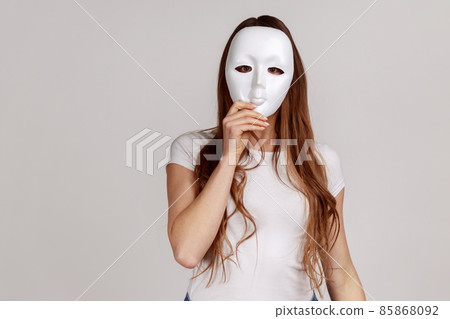 Portrait of anonymous dark haired woman covering her face with white mask, hiding personality, conspiracy and privacy, wearing white T-shirt. Indoor studio shot isolated on gray background. 85868092