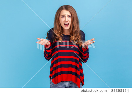 Nervous disappointed woman wearing striped casual style sweater spreading hands and shouting asking what do you want from me. annoyed expression. Indoor studio shot isolated on blue background. 85868154