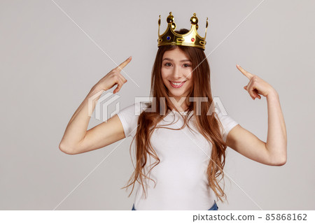 Happy cheerful woman pointing fingers on golden crown on her head, showing her leadership qualities, concept of authority, wearing white T-shirt. Indoor studio shot isolated on gray background. 85868162