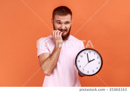 Portrait of anxious nervous bearded man biting nails on his fingers holding big wall clock in hand, worried about deadline, wearing pink T-shirt. Indoor studio shot isolated on orange background. 85868163