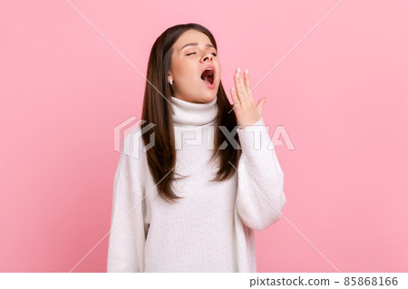 Sleepy young woman standing yawning with closed eyes and covering mouth, feeling exhausted need rest, wearing white casual style sweater. Indoor studio shot isolated on pink background. 85868166