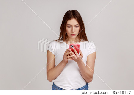 Portrait of concentrated woman using smart phone, typing message or checking social networks, writing post, serious expression, wearing white T-shirt. Indoor studio shot isolated on gray background. 85868167