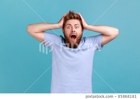 Portrait of pleasant looking cheerful bearded man with mouth open in amazement, expressing shock, astonishment, keeping hands on his head. Indoor studio shot isolated on blue background. 85868181