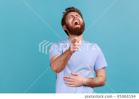 Hilarious laughter. Portrait of joyful happy bearded man laughing loudly and pointing to camera, mocking taunting you, holding belly. Indoor studio shot isolated on blue background. 85868194