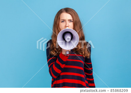 Portrait of woman wearing striped casual style sweater, taking in megaphone, looking at camera with serious expression, important announcement. Indoor studio shot isolated on blue background. 85868209