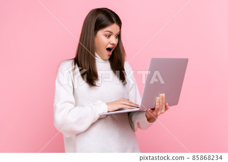 Side view of astonished female working on laptop, being shocked of results of her work, open mouth, wearing white casual style sweater. Indoor studio shot isolated on pink background. 85868234