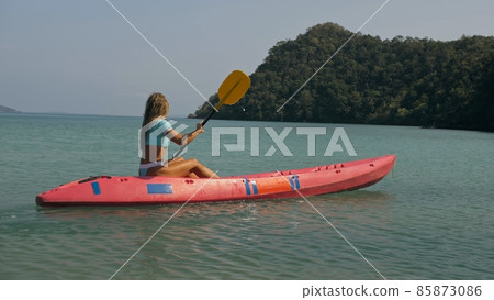 Young blonde woman in blue swimsuit rows pink plastic canoe along azure sea bay past island with palms under blue sky at resort. 85873086