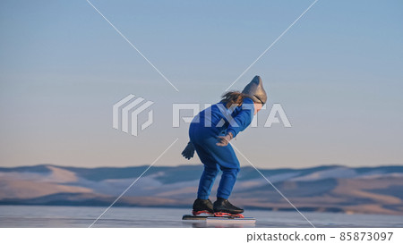 The child train on ice professional speed skating. The girl skates in the winter in sportswear, sport glasses, suit. Outdoor slow motion. 85873097