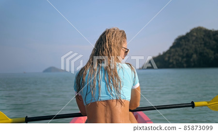 Long curly haired blonde woman in swimsuit rows plastic kayak along azure ocean water at exotic resort close backside view. 85873099