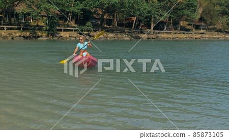 Young blonde woman in blue swimsuit rows pink plastic canoe along azure sea bay past island with palms under blue sky at resort. 85873105