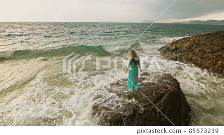 Woman walks on rock of sea reef stone, stormy cloudy ocean. Blue swimsuit dress tunic. Concept rest, tropical resort coastline tourism summer holidays Woman walks on rock of sea reef stone, stormy cloudy ocean. Blue swimsuit dress tunic. Concept rest, tropical resort coastline tourism summer holidays 85873239