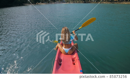 Seductive blonde woman in blue swimsuit sails on pink plastic kayak along azure sea at exotic resort backside upper above view. 85873256