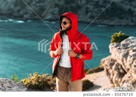 Portrait of a brutal bearded man in a red jacket by the sea. Male portrait on the background of a seascape. A traveler on the background of the sea coast. A man in a jacket at sunset 85876719