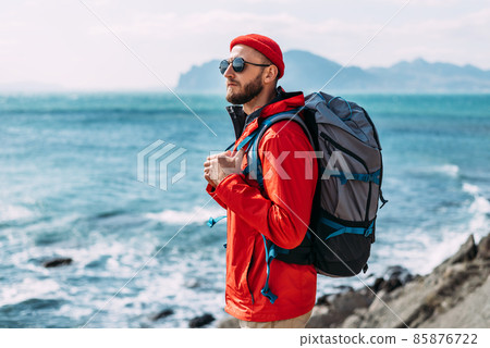 A bearded traveler with a backpack on the background of the sea. Portrait of a traveler in red clothes and sunglasses. A tourist with a backpack stands against the background of the sea. Copy space 85876722