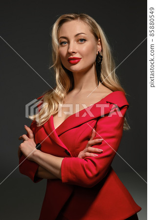 Blonde girl, bright make-up and deep neckline, hands folded, in red dress and black earrings. Smiling, posing sideways on gray background. Close up 85880549