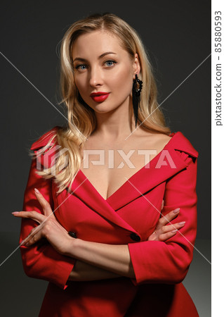 Gorgeous blonde model, bright make-up and deep neckline, crossed hands, in red dress and black earrings. She posing against gray background. Close up 85880593