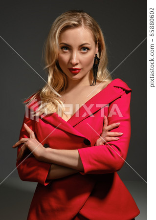 Blonde lady with bright make-up and deep neckline, crossed hands, in red dress and black earrings. She is posing against gray background. Close up 85880602
