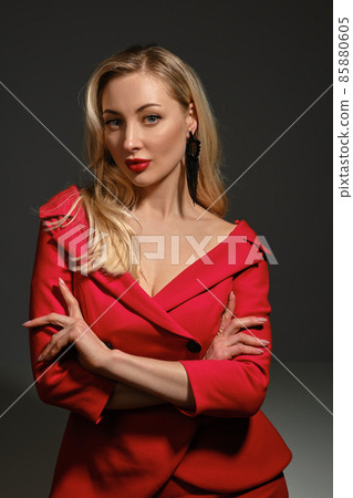 Blonde girl with deep neckline, hands folded, in red dress and black earrings. She looking at you, posing on gray background. Copy space, close up 85880605