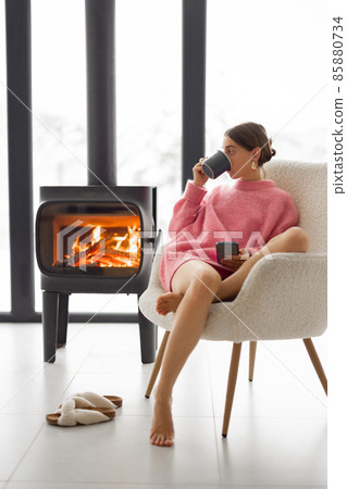 Woman with cup on chair by the fireplace at house on nature Woman with cup on chair by the fireplace at house on nature 85880734