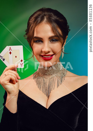 Brunette model in black dress, necklace and earrings. Smiling, showing two playing cards, posing on colorful background. Poker, casino. Close-up Brunette model in black dress, necklace and earrings. Smiling, showing two playing cards, posing on colorful background. Poker, casino. Close-up 85881136