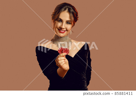 Brunette girl in black dress and shiny jewelry. She smiling, showing two red chips, posing on brown background. Poker, casino. Close-up Brunette girl in black dress and shiny jewelry. She smiling, showing two red chips, posing on brown background. Poker, casino. Close-up 85881411