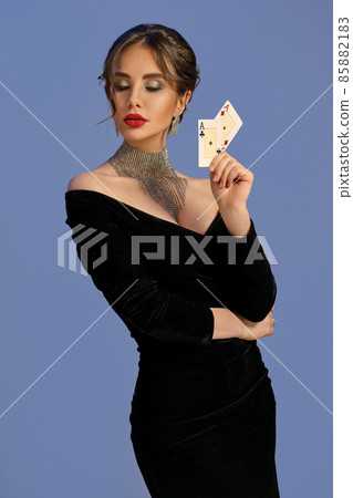 Brunette woman with bare shoulders, in black dress and jewelry. Showing two playing cards, posing on blue background. Poker, casino. Close-up Brunette woman with bare shoulders, in black dress and jewelry. Showing two playing cards, posing on blue background. Poker, casino. Close-up 85882183