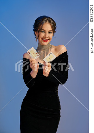 Brunette female with bare shoulders, in black dress and jewelry. Smiling, showing two aces, posing on blue background. Poker, casino. Close-up 85882201