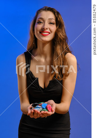 Brunette lady in black dress. She is smiling, showing handful of colorful chips, posing on blue background. Poker, casino. Close-up Brunette lady in black dress. She is smiling, showing handful of colorful chips, posing on blue background. Poker, casino. Close-up 85882479