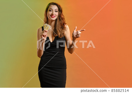 Girl in black dress. Holding two playing cards, smiling, pointing at something, posing colorful background. Poker, casino. Template, mockup. Close up 85882514