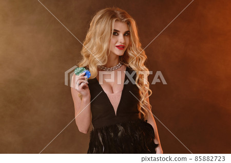 Blonde woman in black dress and necklace. Smiling, showing green and blue chips, posing on dark smoky studio background. Poker, casino. Close-up 85882723