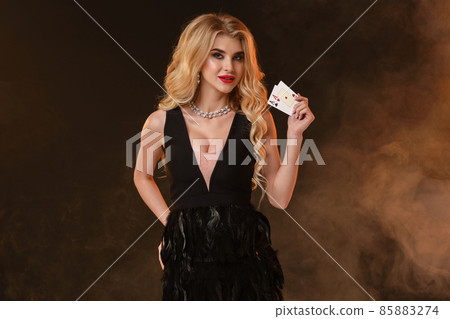 Blonde lady in black dress with feathers and necklace. She is smiling, showing two aces, posing on black smoky background. Poker, casino. Close-up Blonde lady in black dress with feathers and necklace. She is smiling, showing two aces, posing on black smoky background. Poker, casino. Close-up 85883274